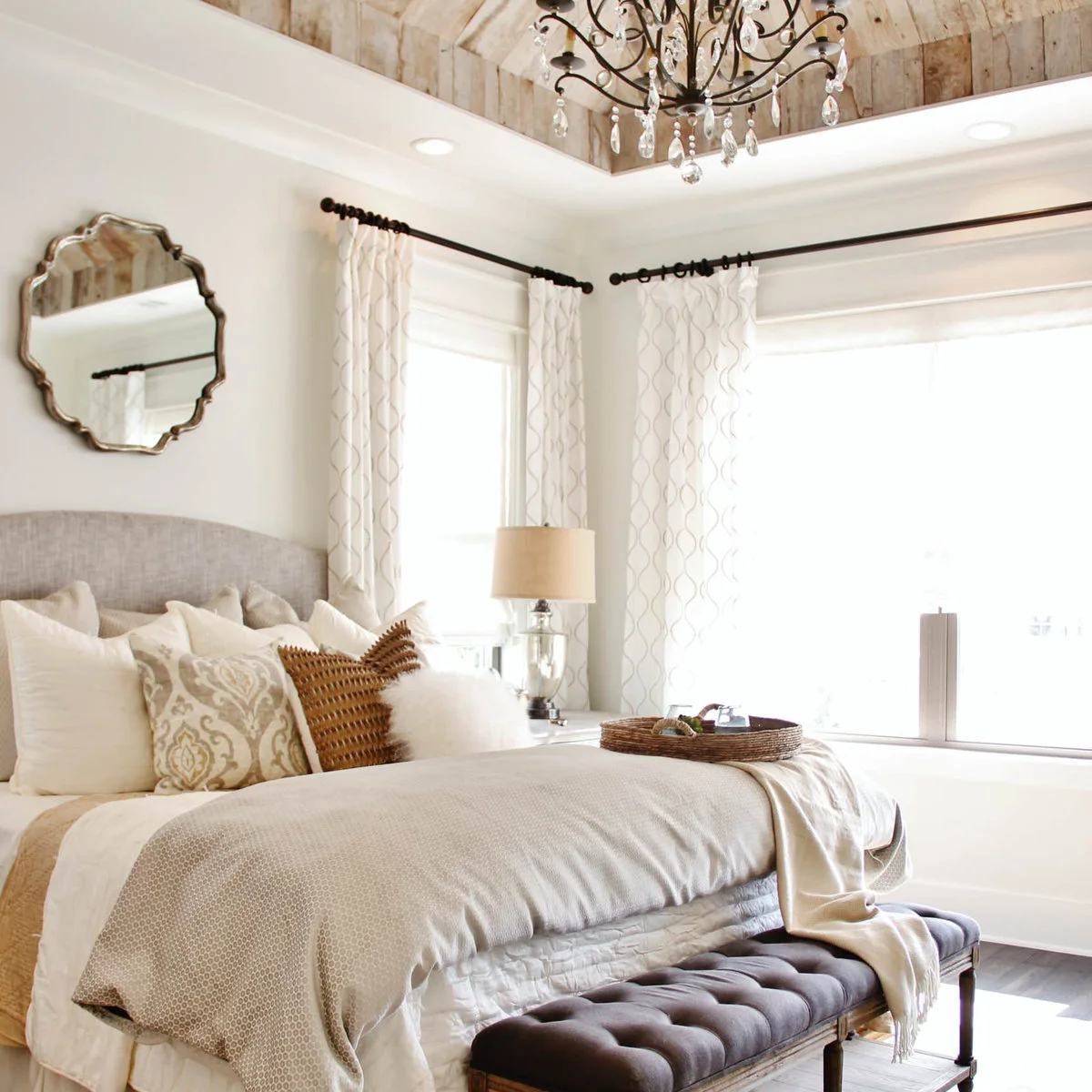 Pale bedroom with a curved gray headboard, dark tufted bench and light patterned curtains