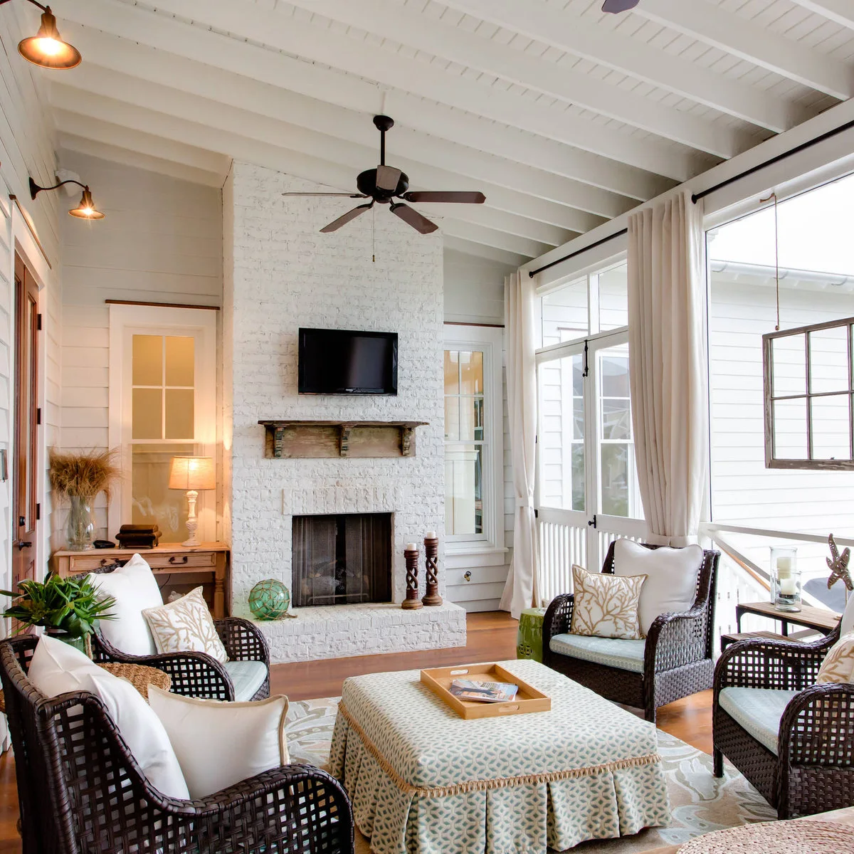 Dark woven chairs and a sofa surround a large patterned ottoman in a white porch with a brick fireplace wall.