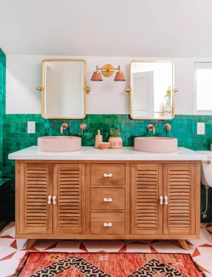 Two pink bowl basins sit on a timber vanity in front of emerald green tiles beneath warm metal-framed mirrors.