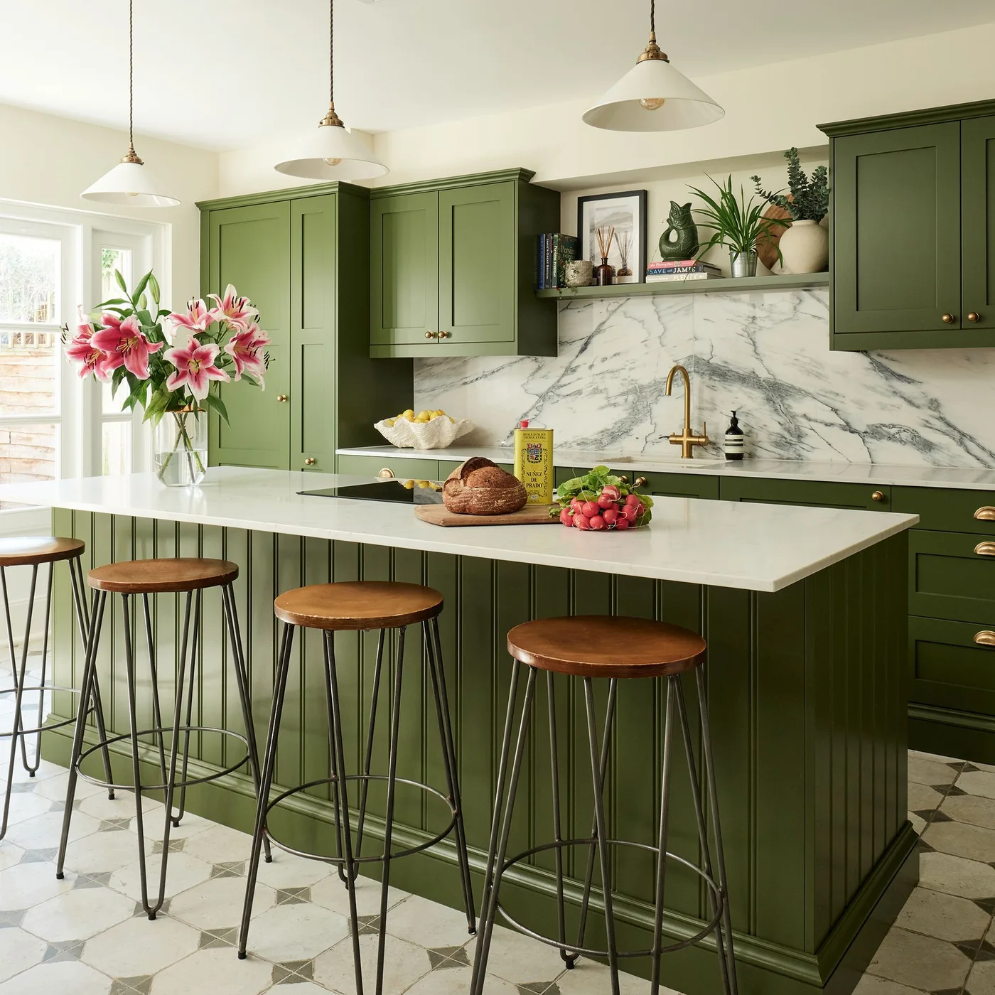 Olive green cabinets and a paneled island surround pale counters and a veined backsplash beneath pale pendant lights