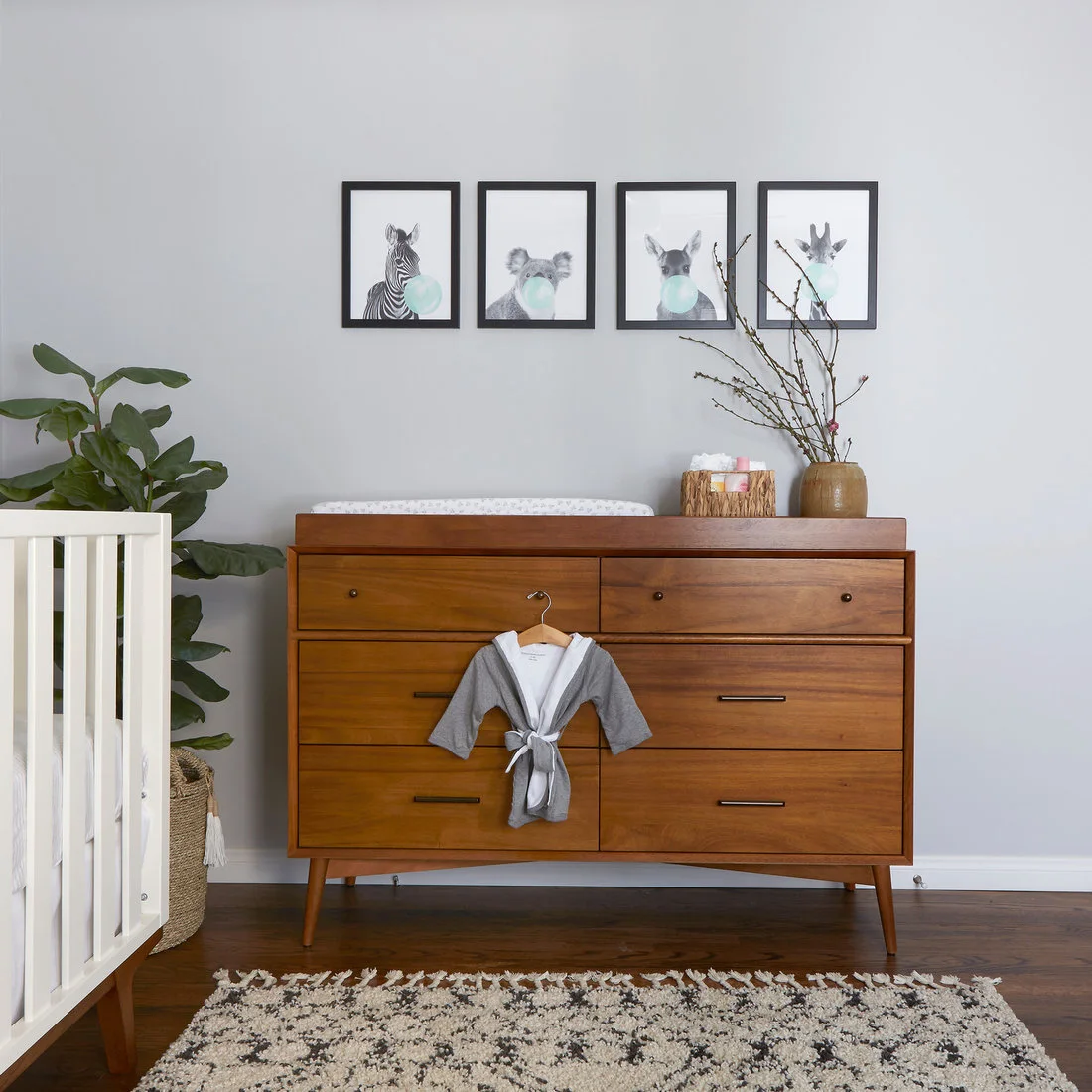 A wood dresser with a changing pad stands against a gray wall below four animal pictures, beside a plant and woven container.