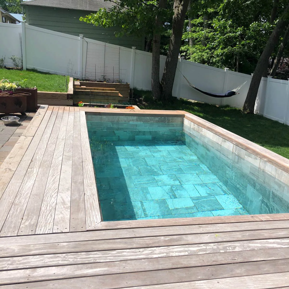 A small rectangular plunge pool sits within a wood deck beside pale paving and garden planting.