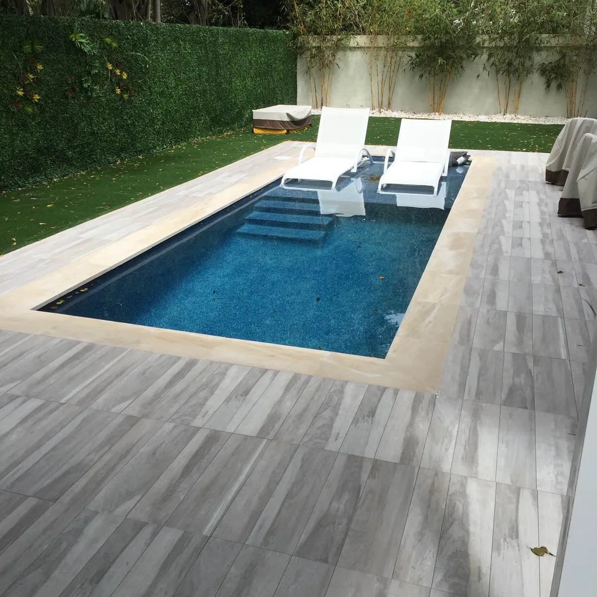 A compact blue rectangular pool has a pale shallow ledge, gray surrounding paving and two white loungers beside a hedge.