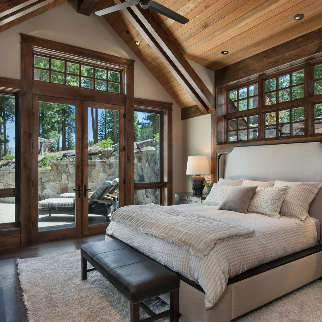 A pale upholstered bed and dark bench beneath timber roof beams beside glazed double doors