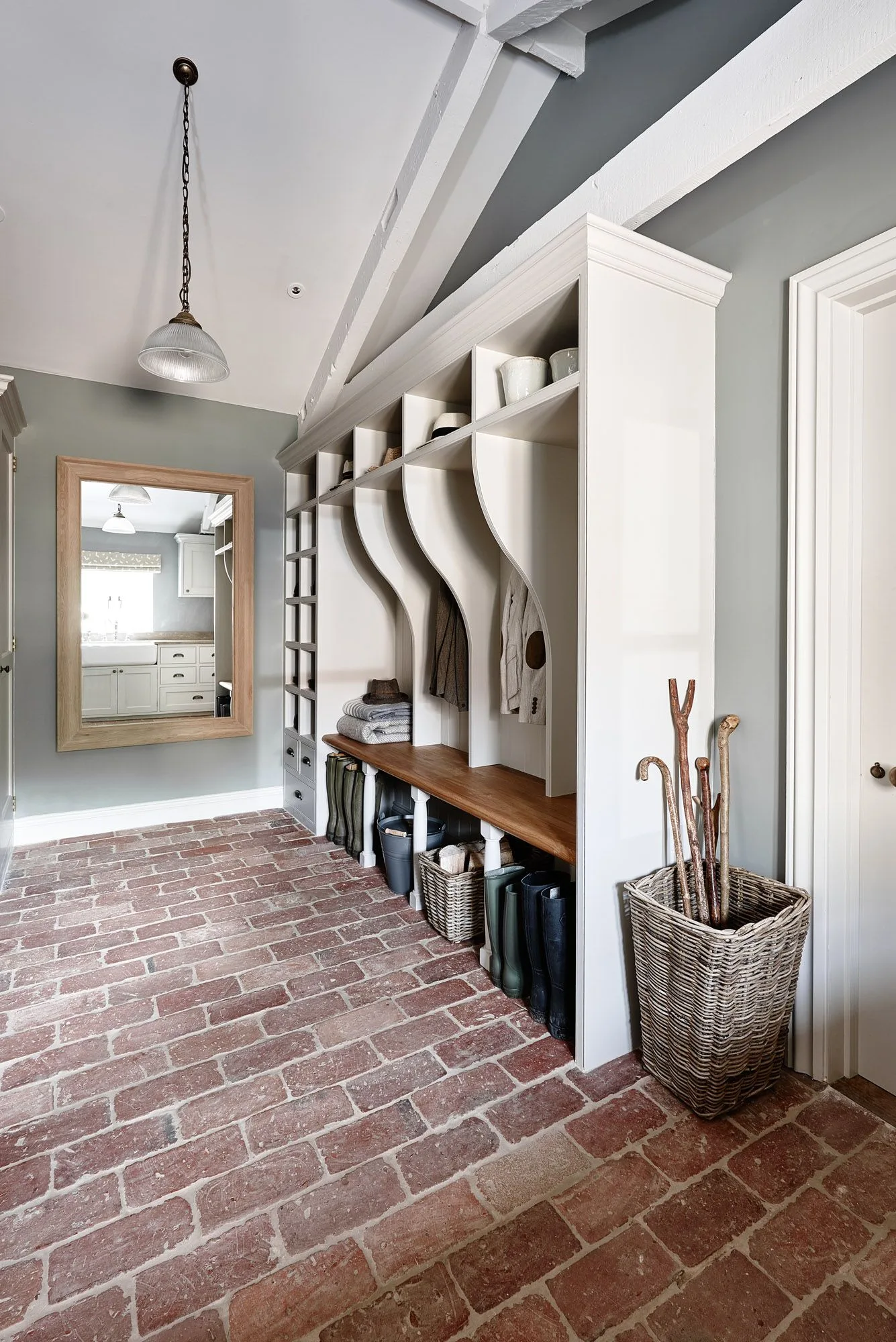 White mudroom storage with curved coat-bay dividers, a wooden bench, gray walls and red brick-toned flooring.