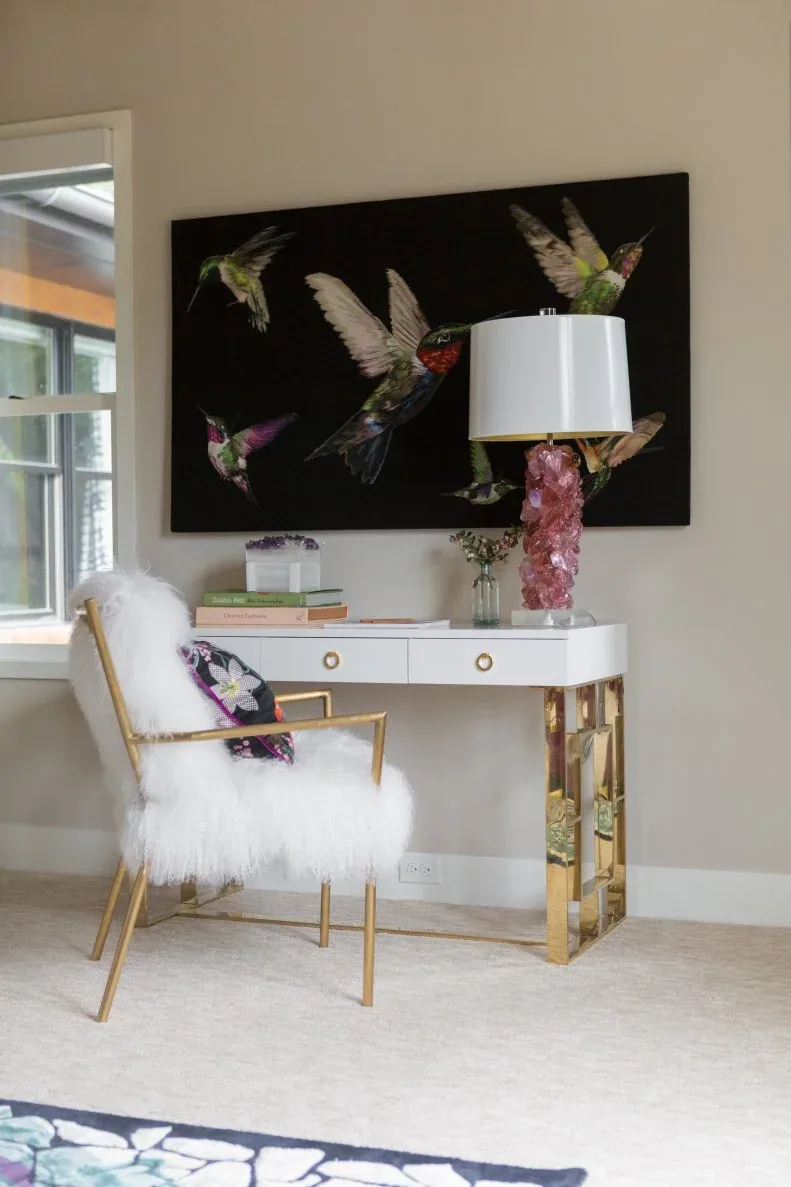 White writing desk with gold-toned legs and drawer rings beside a fluffy pale chair and pink lamp.