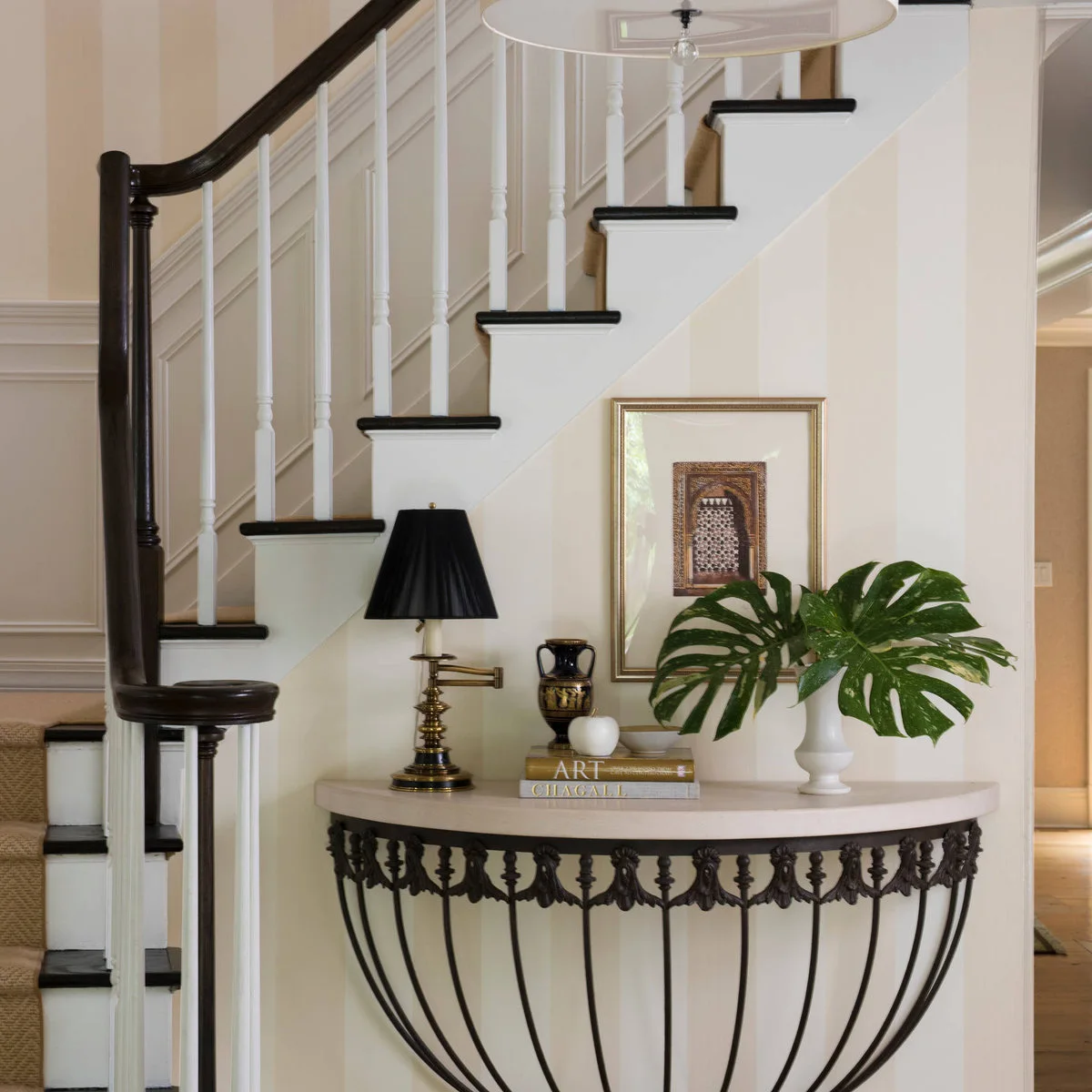 A half-round console beside stairs with a dark-shaded lamp, books and a white vase of large green leaves.