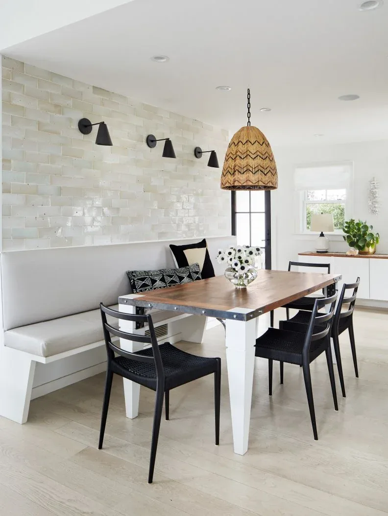 Dining nook with a glossy pale staggered tile wall above a white bench, three black wall lights, wooden table, black chairs, and woven pendant.