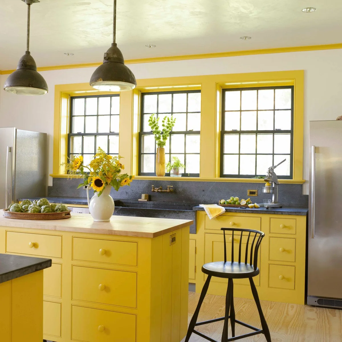 Yellow lower cabinets and window trim sit beside dark counters, dark window frames and pale walls in a bright kitchen.