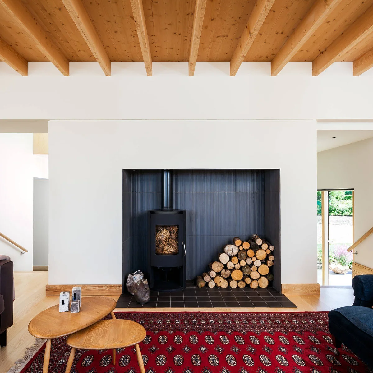 A black curved-front wood stove stands in a broad dark tiled recess below pale walls and exposed timber ceiling beams.