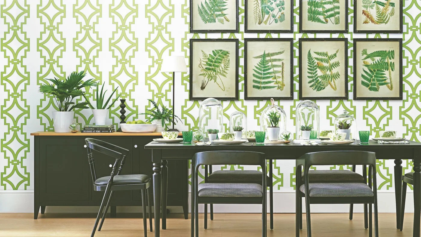 Green geometric wallpaper and framed botanical pictures surround a dark dining table, chairs and sideboard