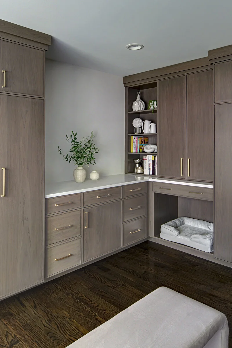 Gray-brown mudroom cabinets turn a corner around a pale countertop, open shelves and a pet-bed recess beneath a drawer.