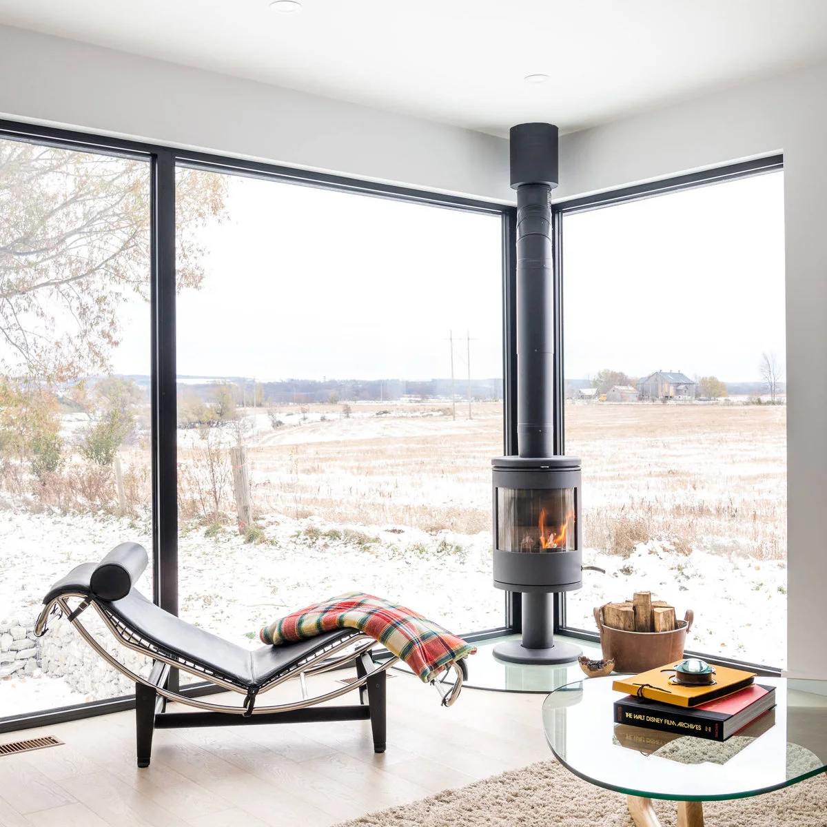 A slim cylindrical stove stands on a curved transparent floor panel beside large corner windows, a reclining chair and a glass table.