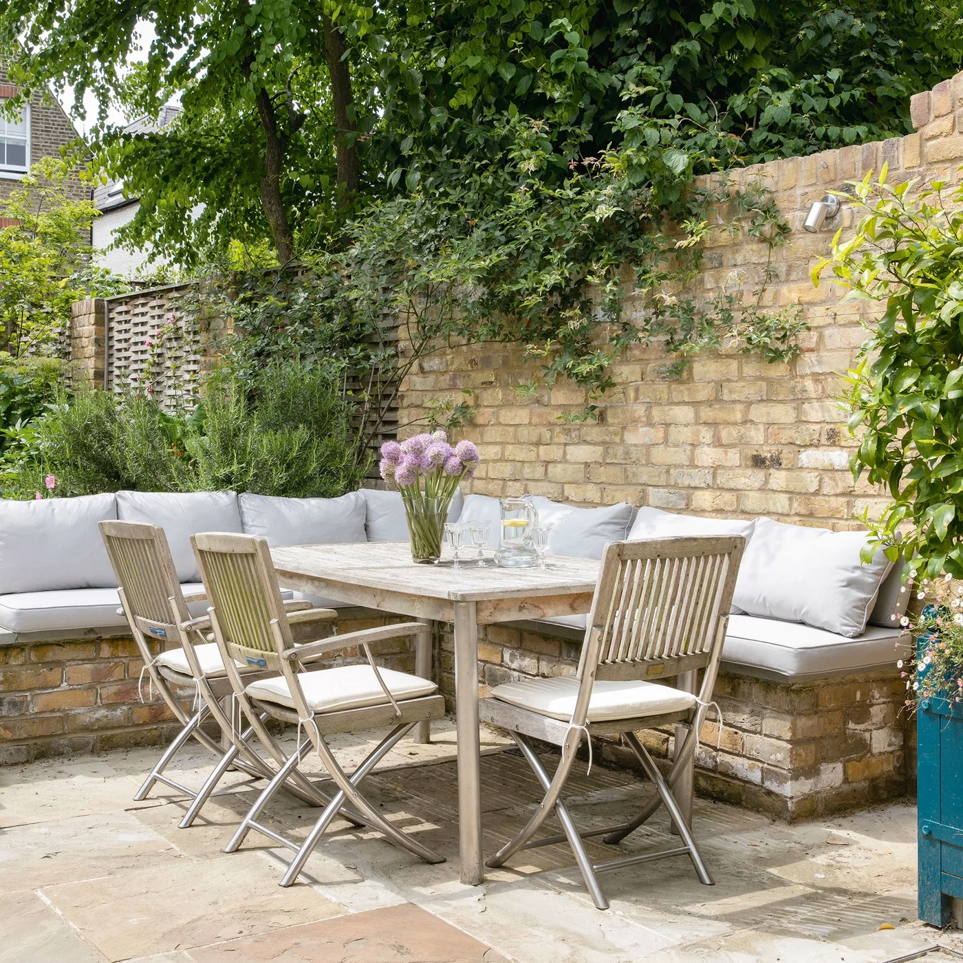 A pale cushioned bench follows two courtyard walls behind a timber dining table and folding chairs