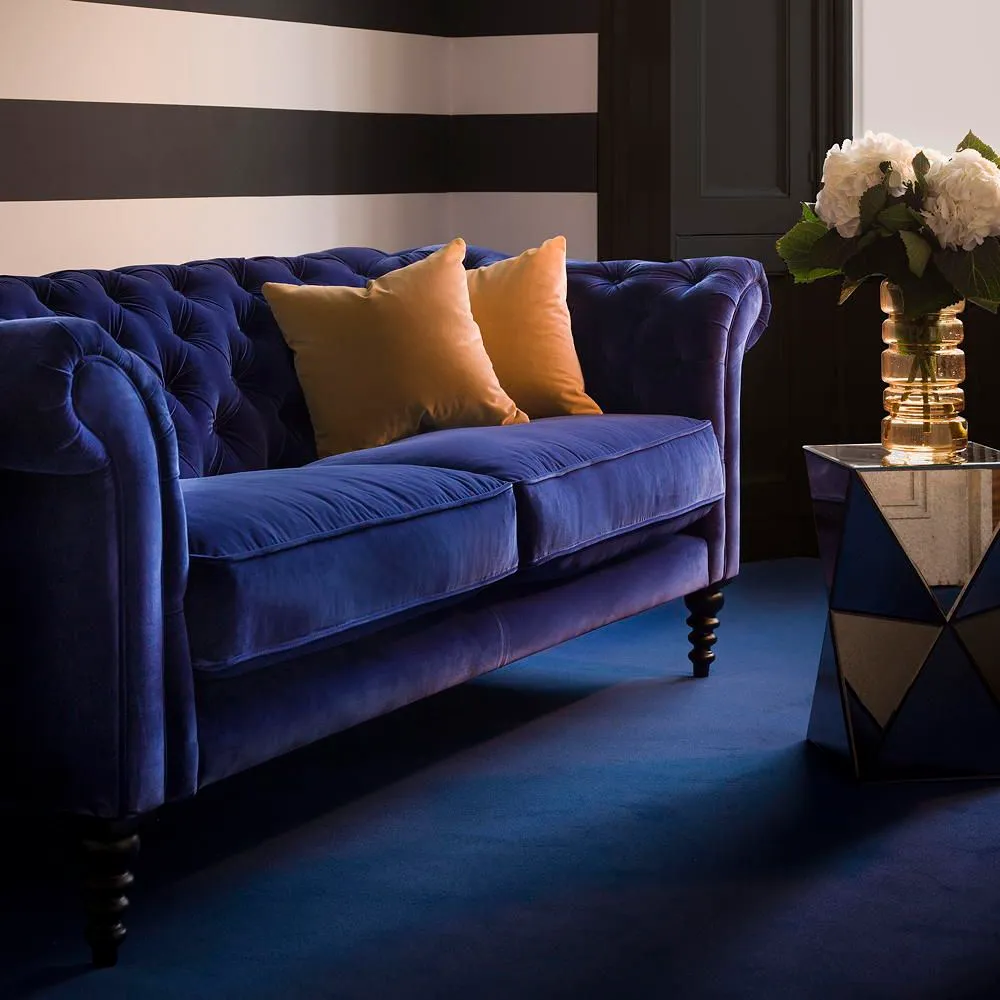 A deep blue tufted sofa sits beside a small dark table with a warm gold cushion.