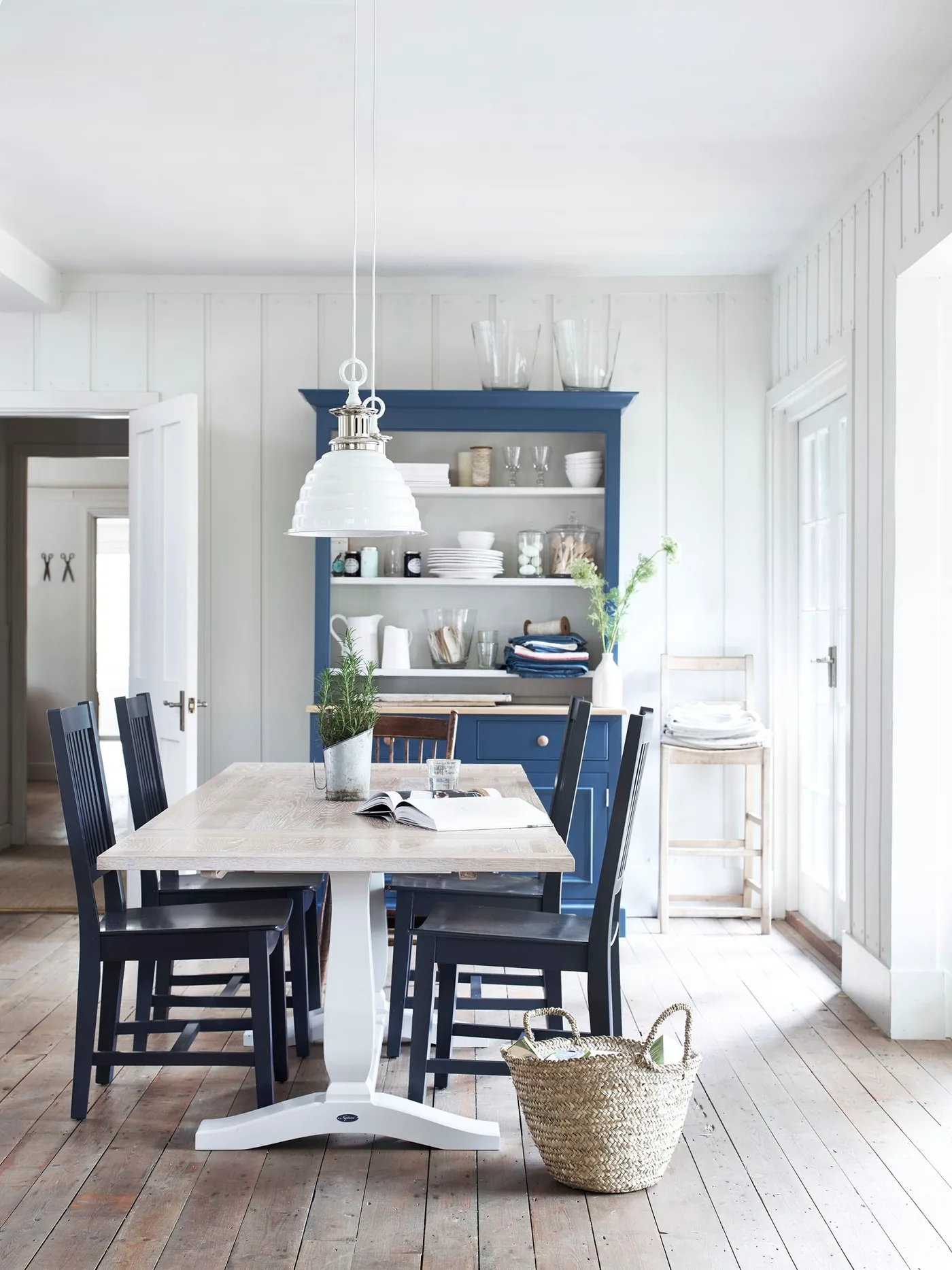 Country dining room with pale paneling, blue dresser displaying tableware, blue chairs, a lighter wooden table and a white pendant light.