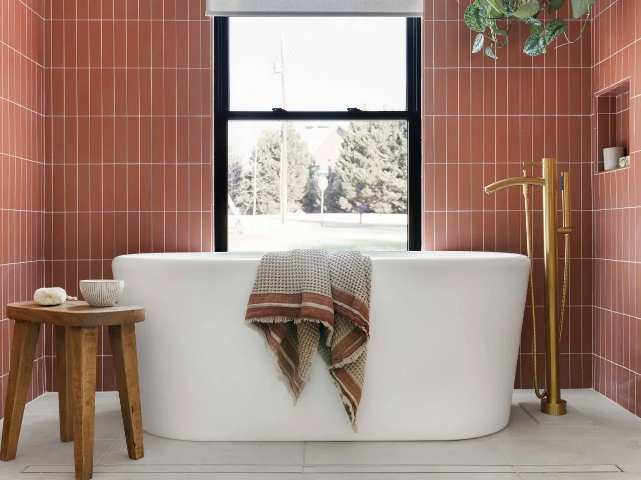Rust-pink vertical tiles surround a white tub beside a timber stool, patterned towel and hanging greenery.