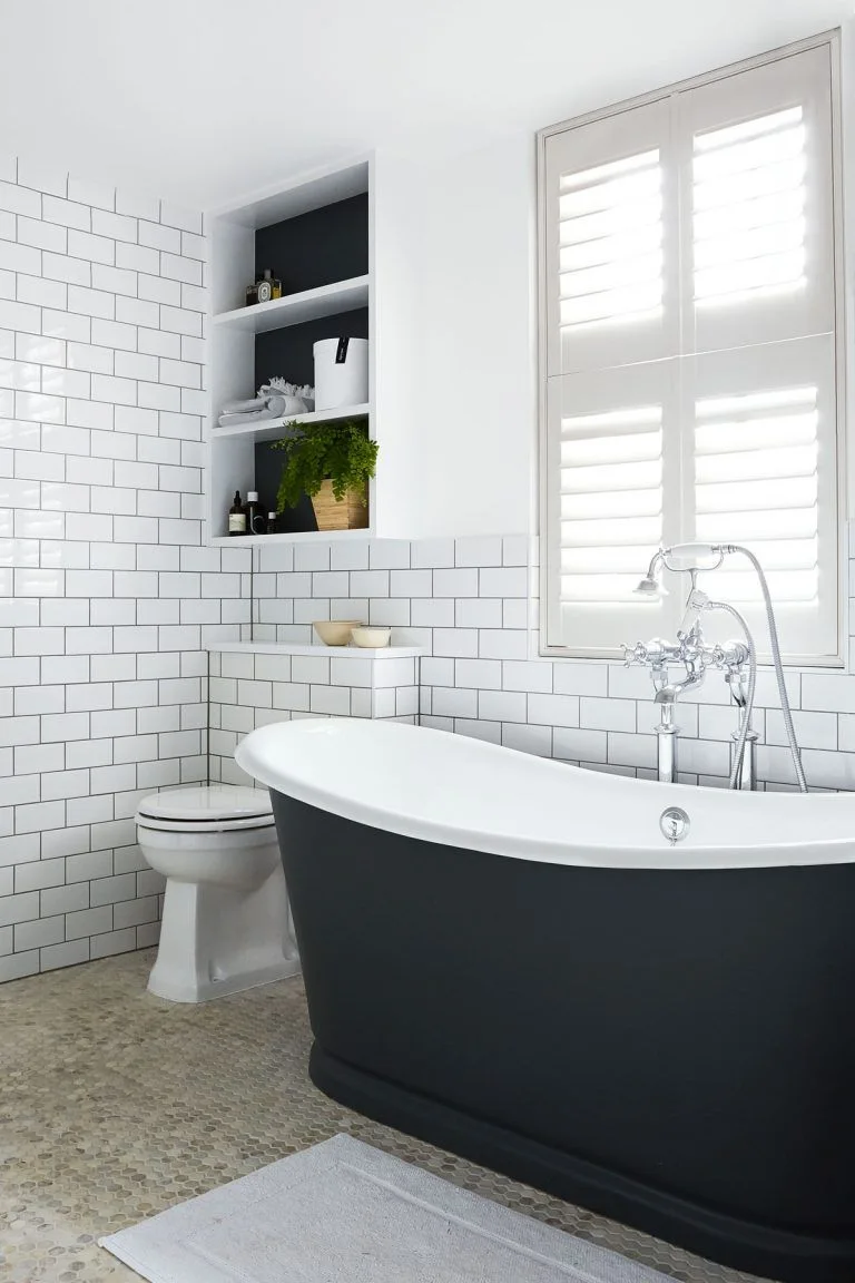 A dark freestanding tub with a white interior sits beside white subway tiles, window shutters, recessed shelving and a pale small-scale floor
