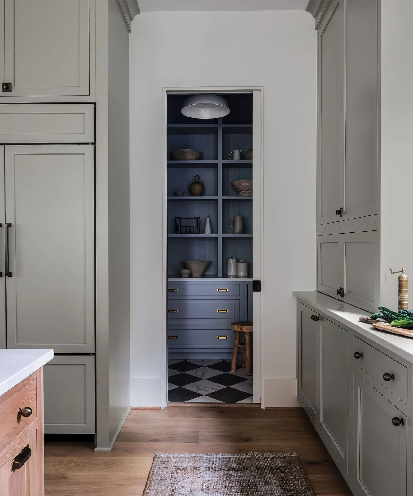 A deep blue pantry with open shelves and lower drawers sits beyond a pale cabinet-lined kitchen with a patterned floor and small hanging light