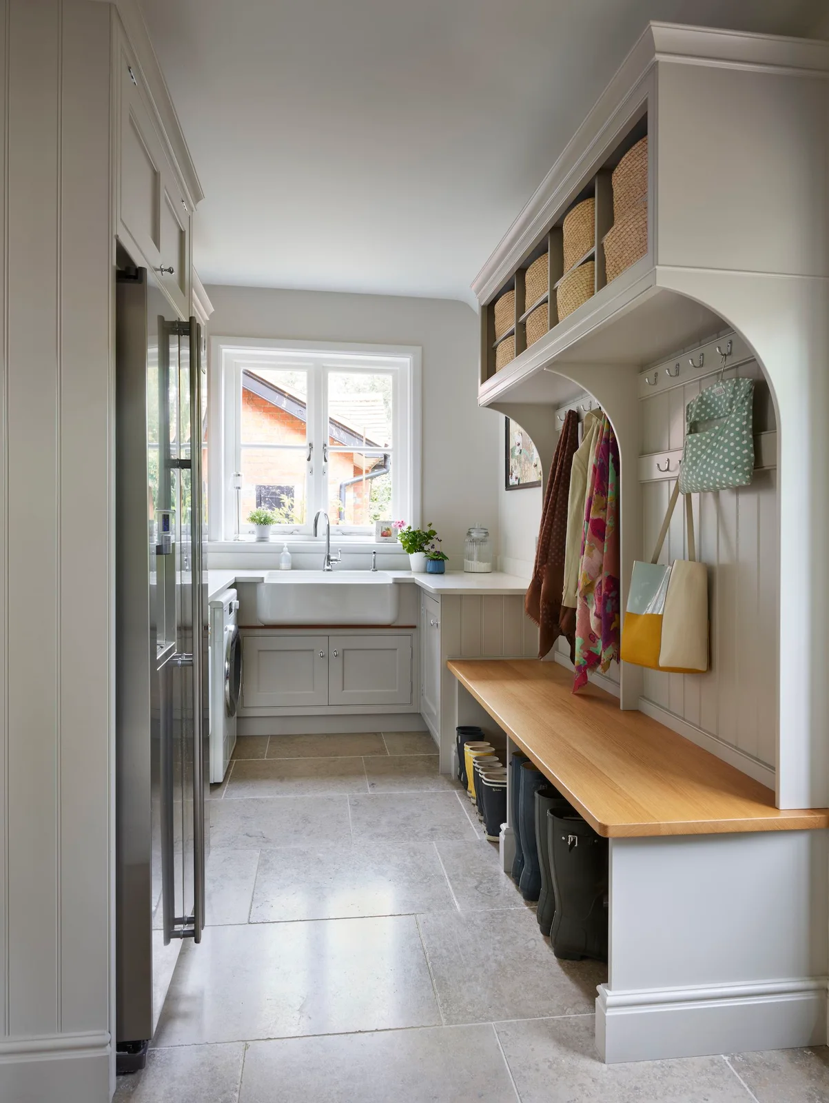 A narrow pale utility room has a long wood bench with boots underneath, overhead baskets and hooks, laundry equipment opposite and a sink beneath the window.