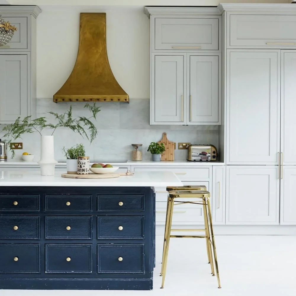Deep navy island drawers sit below a pale counter beside light wall cupboards, a warm-colored hood and a slender metallic stool