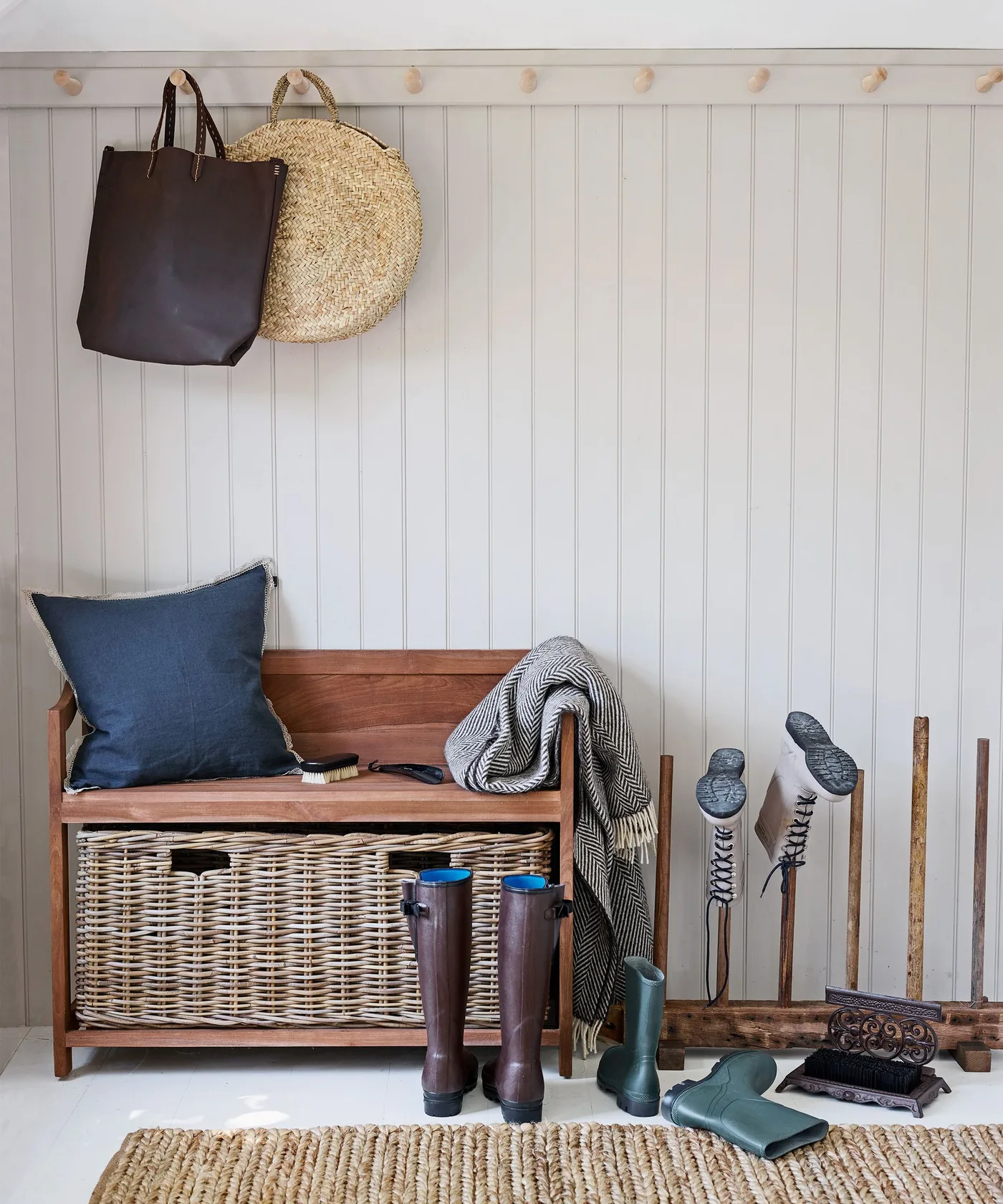 Wood entry bench with a deep woven basket below, blue cushion, wall pegs, boots and a nearby boot rack