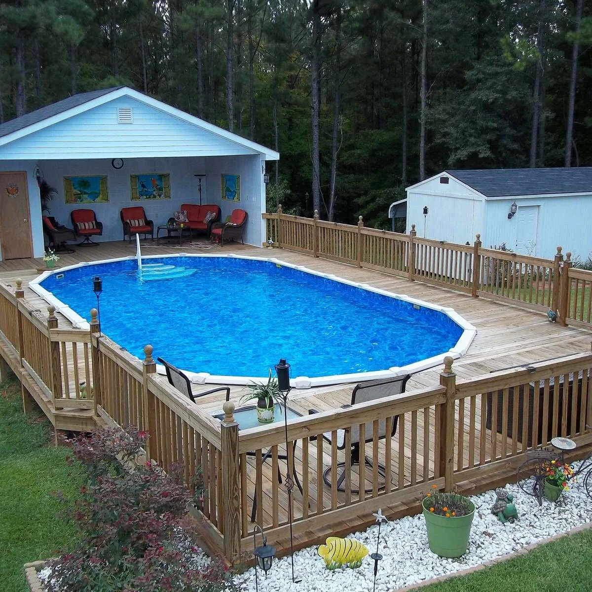 Oval above-ground pool within a wood deck beside a covered sitting area with red cushions and a planted gravel border.