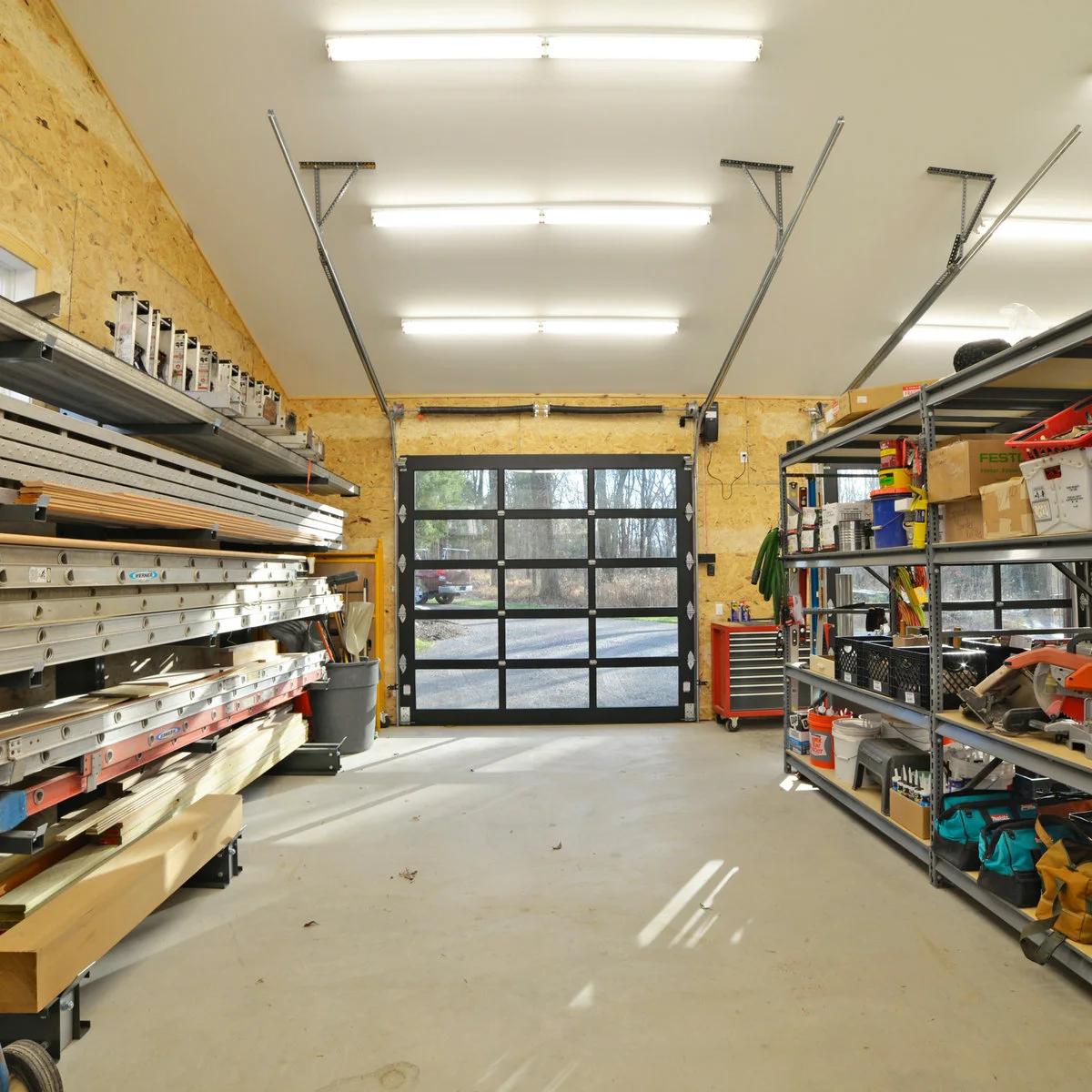 Workshop storage aisle with ladders and long boards on wall racks, open shelving opposite and a glazed garage door at the end.