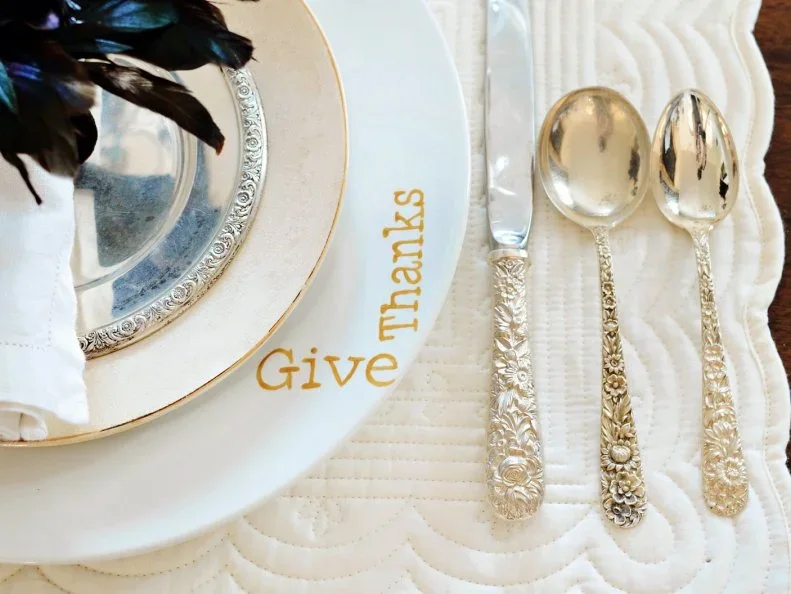 Gold-colored Give Thanks lettering appears on a pale napkin beside silver-colored cutlery and pale plates with warm metallic rims