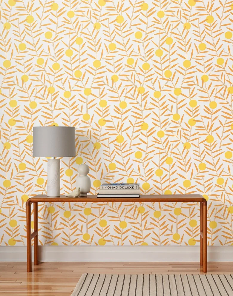 Yellow and ochre leafy wallpaper sits behind a slender wood console with a gray-shaded lamp, books and pale objects above a striped rug.