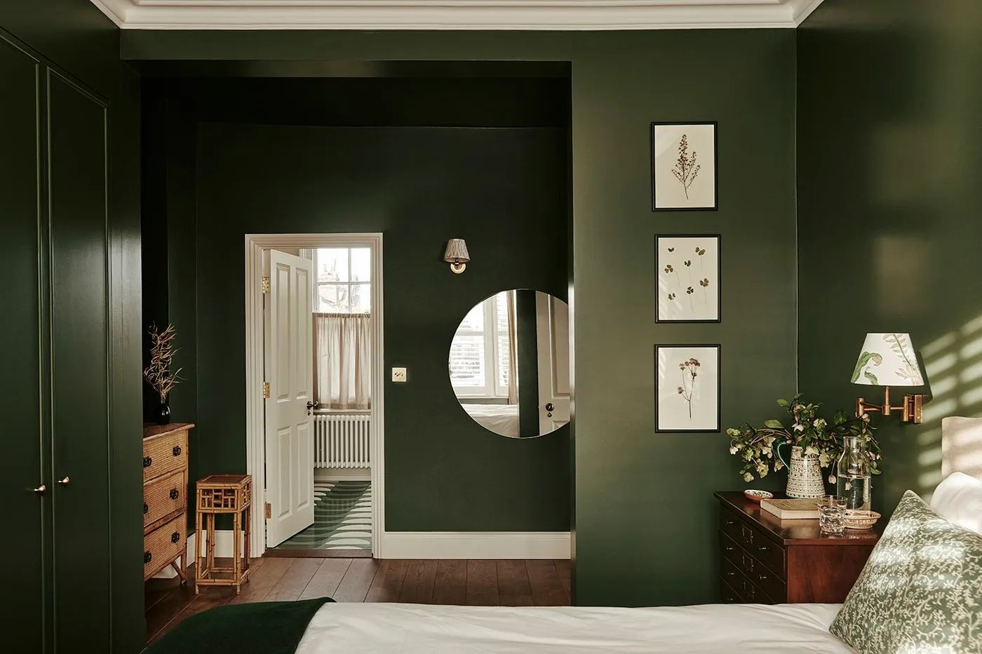 Forest green bedroom with matching wardrobe doors, white bedding, a wood drawer chest, three botanical pictures and a round mirror