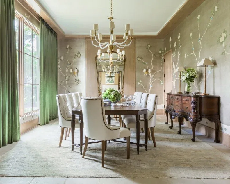 Pale upholstered chairs around a wooden table with flowering wall decoration, green curtains and a carved sideboard.