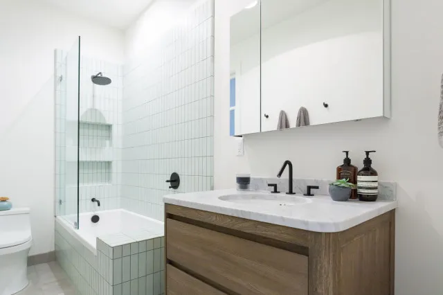 A timber-front vanity sits beneath a broad mirror beside a pale tiled shower.