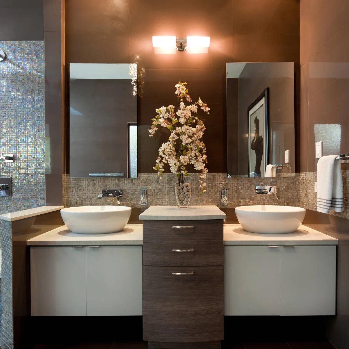 Two white vessel bowls sit on lower pale counters around a taller central wood drawer unit beneath separate mirrors and mosaic tile.