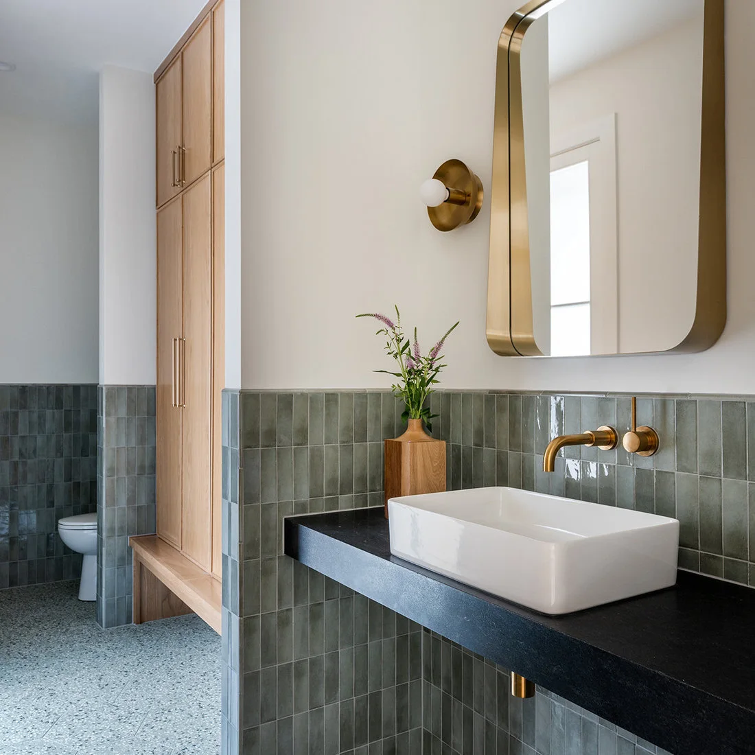 Fine gray terrazzo flooring runs below muted green wall tiles, a white vessel basin and tall timber storage.