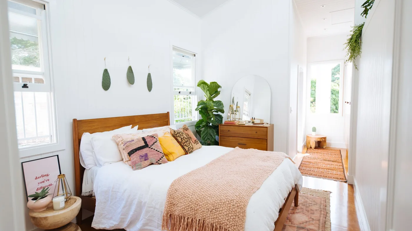 A simple wood bed with white bedding and a pale pink throw stands beside a matching chest, small round bedside table and green plants against white walls