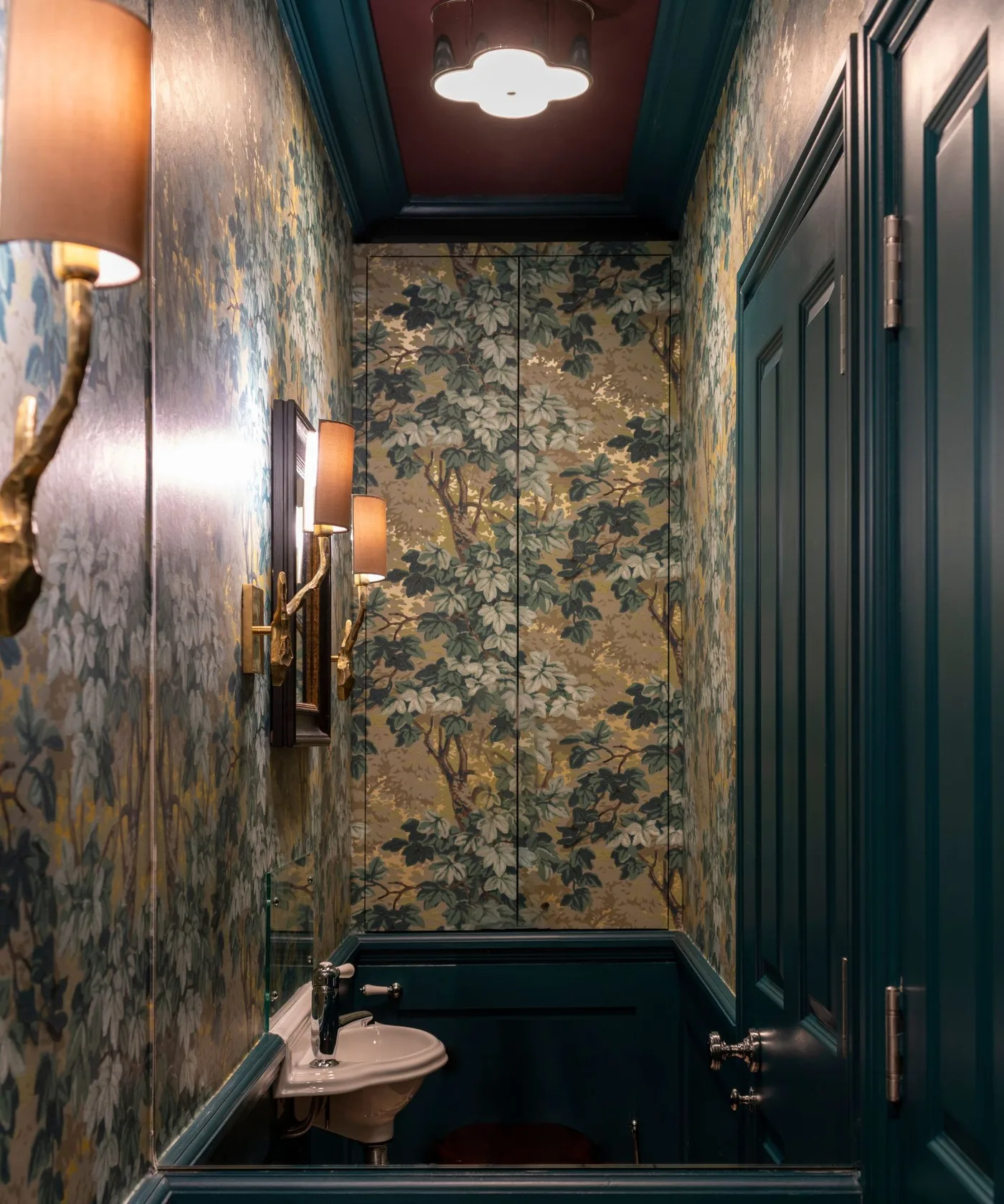 Golden botanical wallpaper fills a narrow powder room beside dark teal paneling, a paneled door, shaded wall lights and a reddish ceiling