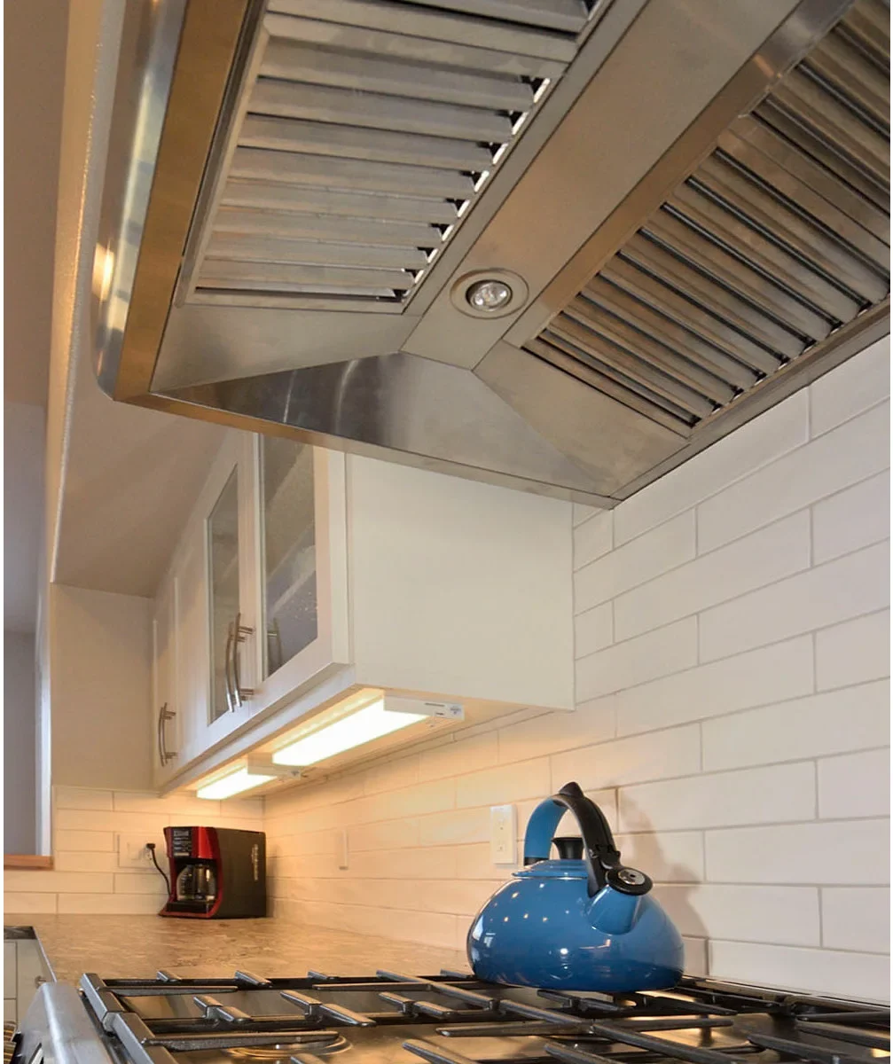 A small round light is visible within a metal range hood beside illuminated under-cabinet fittings above white tile and a blue kettle.
