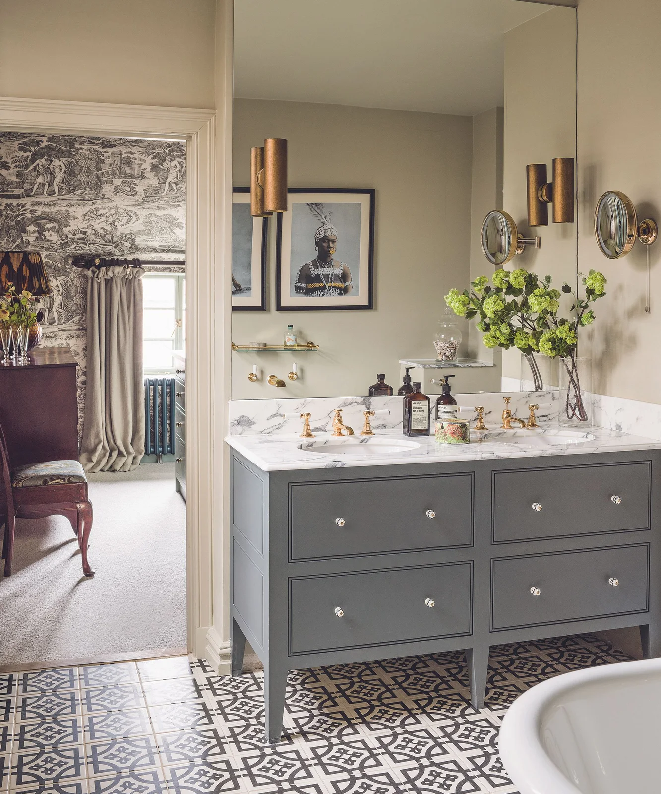 A gray double vanity stands above a patterned monochrome floor beside a mirrored wall and an open doorway to a wallpapered bedroom