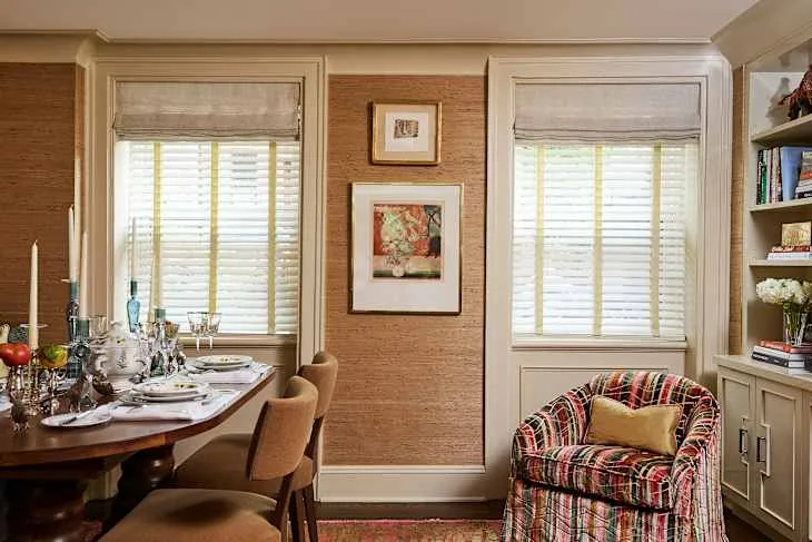 Pale folded fabric treatments above white window slats in a warm-toned dining room with a plaid chair