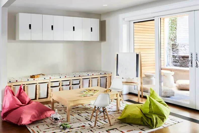 A pale playroom has a long cushioned storage bench with white bins, high white cupboards, a small wood table, bright beanbags and glazed garden doors.