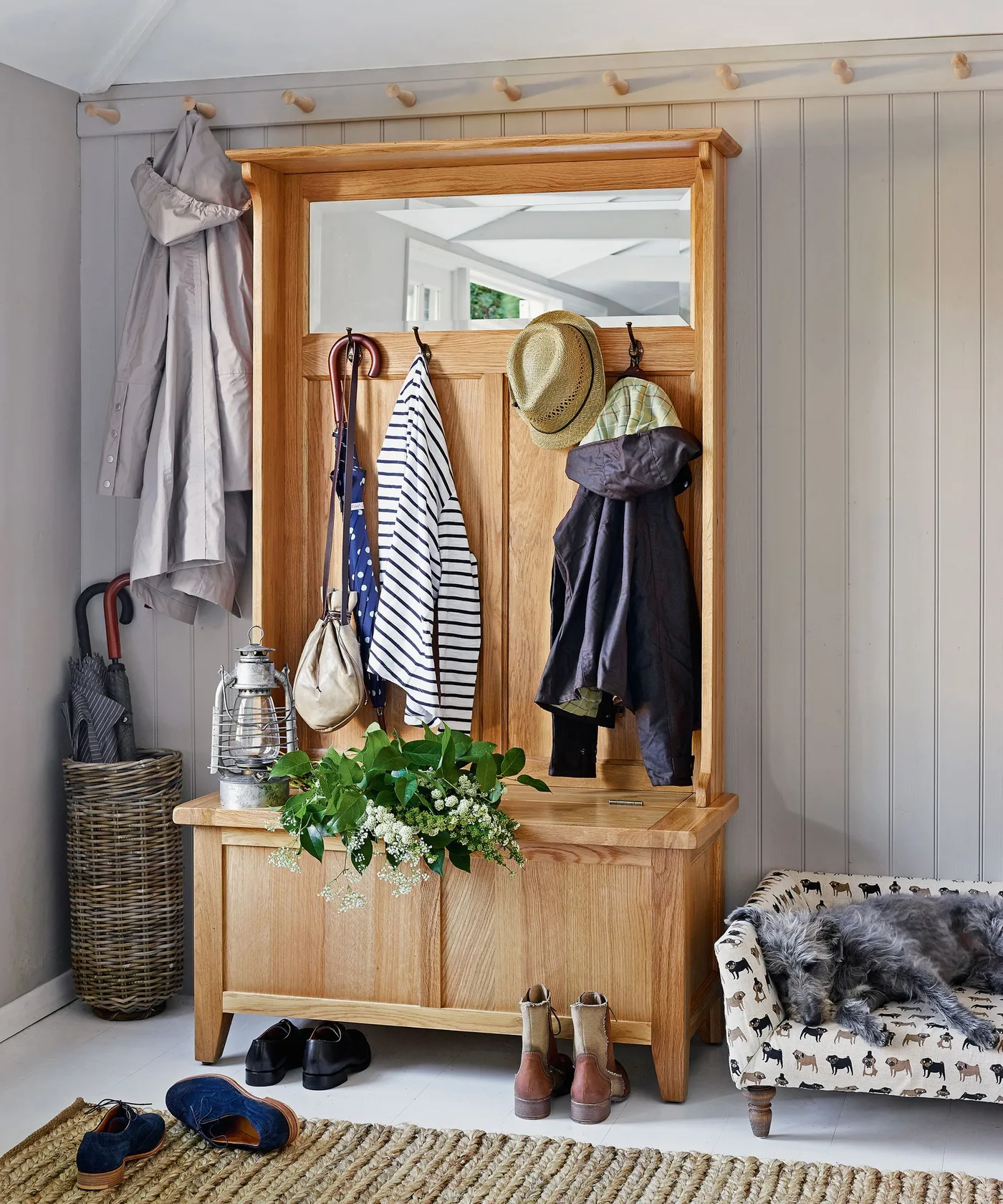Wood hall tree with mirror, hanging hats and coats, a lower storage chest, nearby umbrella basket and a dog bed
