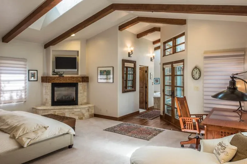 Bedroom with a pale corner fireplace, wood ceiling beams, bed, desk and seating near tall windows.