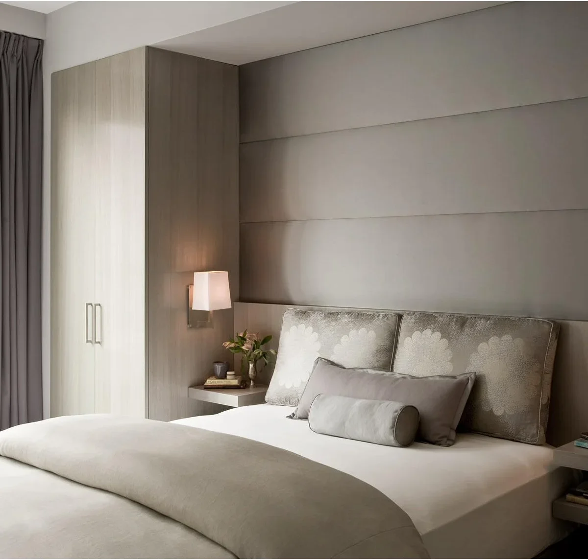 A small white angular wall light is mounted on the side of a wood wardrobe beside a padded bed and floating bedside shelf.