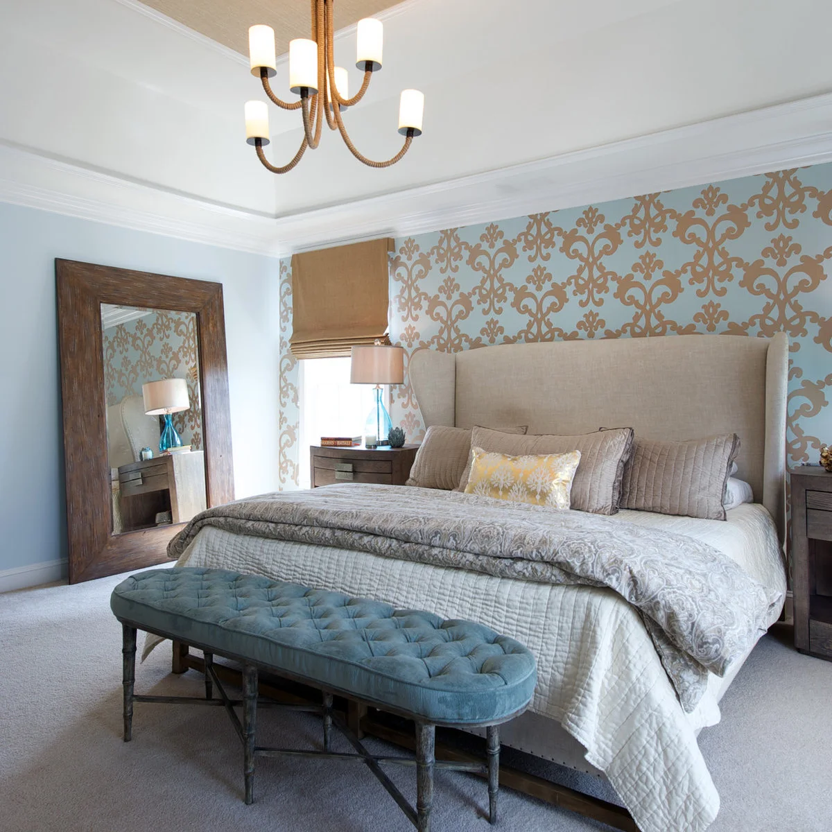 Blue and tan damask wallpaper behind a tall pale headboard with blue bedside lamps and a blue bench.