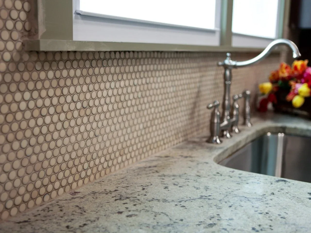 Pale round mosaic tiles with darker grout sit behind a kitchen sink beneath a window.