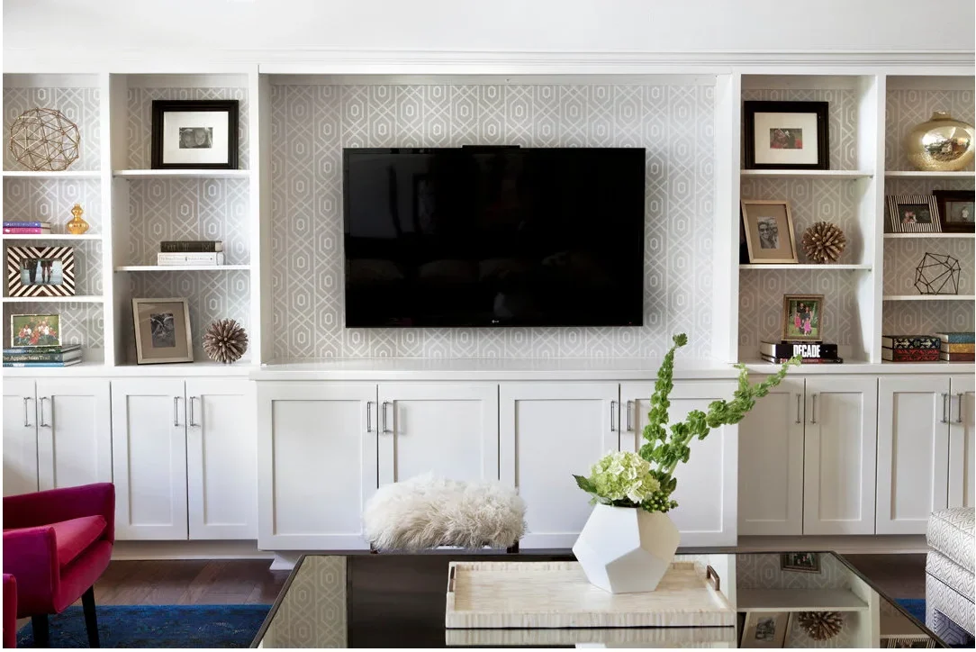 Pale geometric wallpaper runs behind a television and white shelves above closed cupboards.