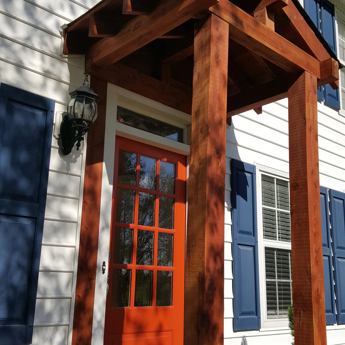 Compact wood-colored front portico framing an orange door beside white siding and blue shutters