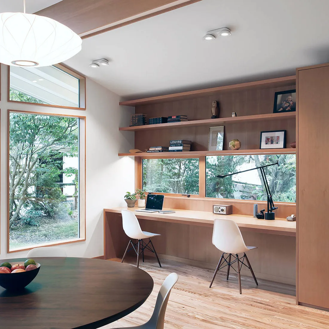 Round dark dining table beside a long wood desk with two white chairs and windows