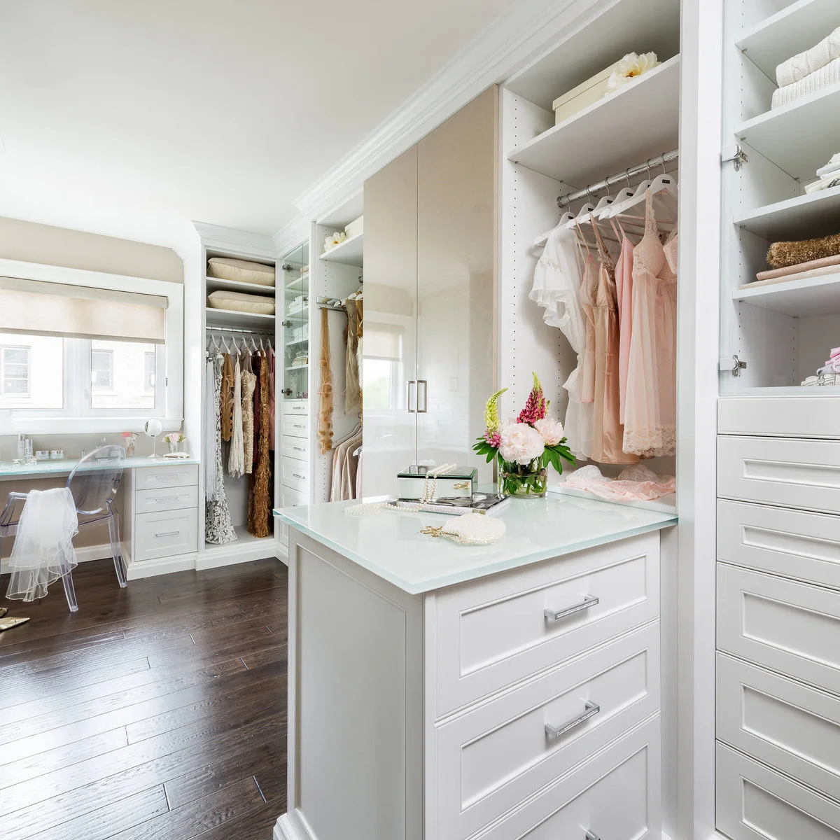 A small vanity beneath a window sits at the end of a fitted dressing room with pale cabinets